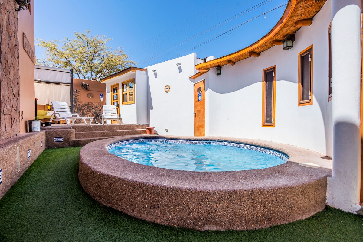 A round, inviting hot tub is situated within a private outdoor area, surrounded by low, rounded walls. Sun loungers are positioned nearby on grassy turf, and bright blue skies are visible overhead, enhancing the tranquil atmosphere.