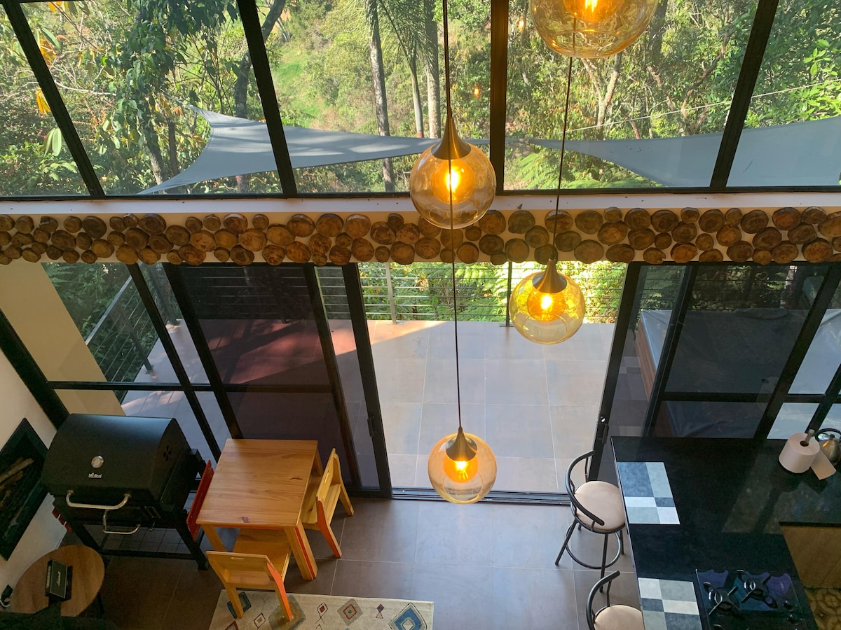 The image captures a spacious interior area with large glass panels that provide views of the surrounding greenery. Decorative light fixtures hang from the ceiling, illuminating a dining table accompanied by chairs. An outdoor area is visible through the glass doors, enhancing the connection with nature.