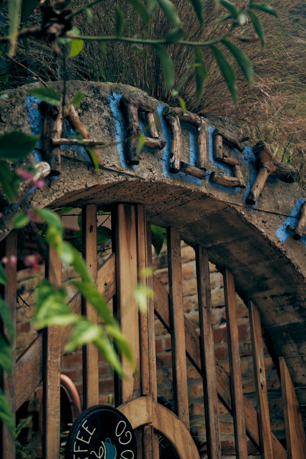 An archway made of stone and wood features the name of the café in blue lettering. Lush greenery surrounds the entrance, creating a natural frame. A wooden sign with the café's logo is visible just below the arch, indicating the welcoming atmosphere of the space.