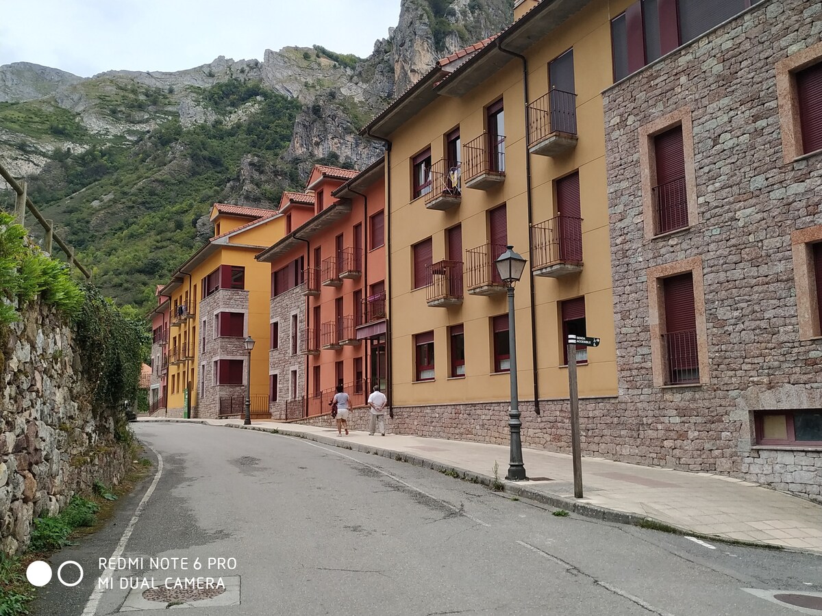 A quiet street in Pola de Somiedo is lined with colorful buildings, characterized by a mix of stone and vibrant painted facades. Lush greenery and mountainous terrain rise in the background, while pedestrians walk along the smooth sidewalk, offering a sense of the tranquil atmosphere.