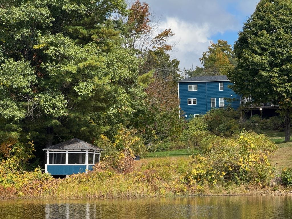A serene lakefront view showcases a blue two-story townhouse nestled among trees with vibrant autumn foliage. A gazebo with a shingled roof is positioned near the water's edge, blending harmoniously with the natural surroundings.