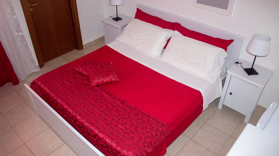 A spacious bed is placed against a textured white wall, featuring red and white bedding. Beside the bed, two nightstands with lamps provide soft lighting. A wooden door is visible in the background, indicating access to the room.