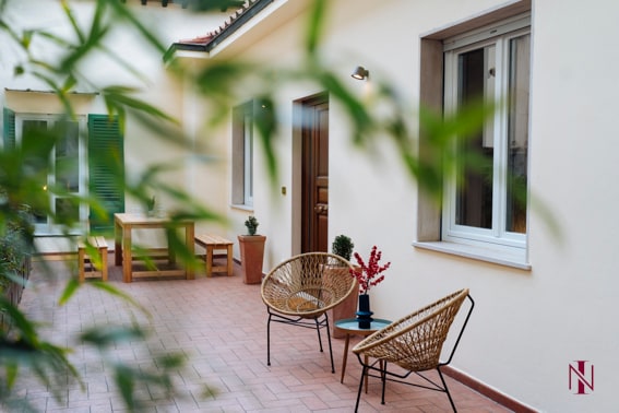 An outdoor terrace is featured with two wicker chairs arranged beside a wooden table. Potted plants add greenery to the space, while a door leads into the apartment. Light illuminates the area, highlighting the neutral tones of the walls.