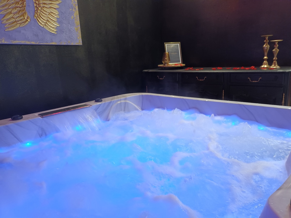 A spa hot tub is surrounded by a dark wall, featuring glowing blue water with bubbling jets. Gold accents, including candlesticks and a framed picture, provide an elegant touch to the space, while rose petals are scattered nearby for a romantic atmosphere.
