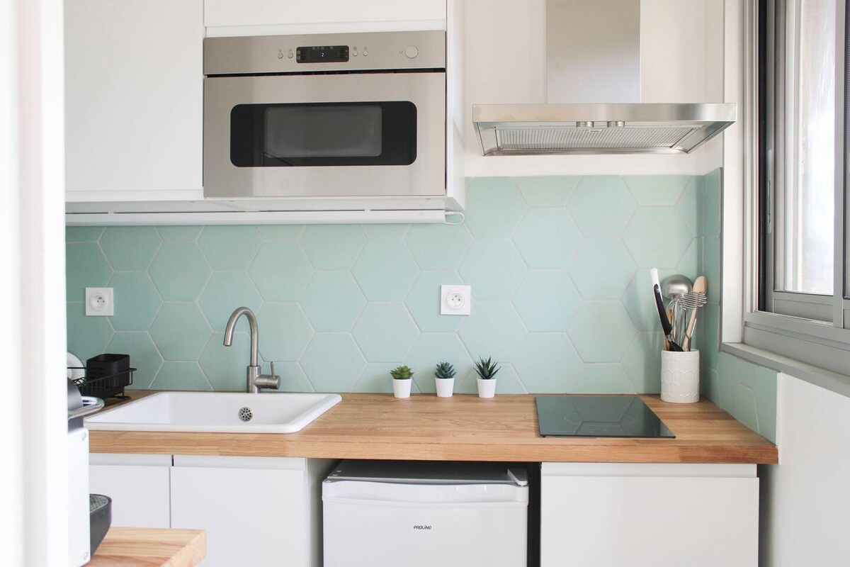 A compact kitchen area features modern appliances, including a microwave and a stovetop. Light wooden countertops complement a mint green tiled backsplash. Small potted plants and kitchen utensils are arranged neatly on the counter, enhancing the functionality of the space.