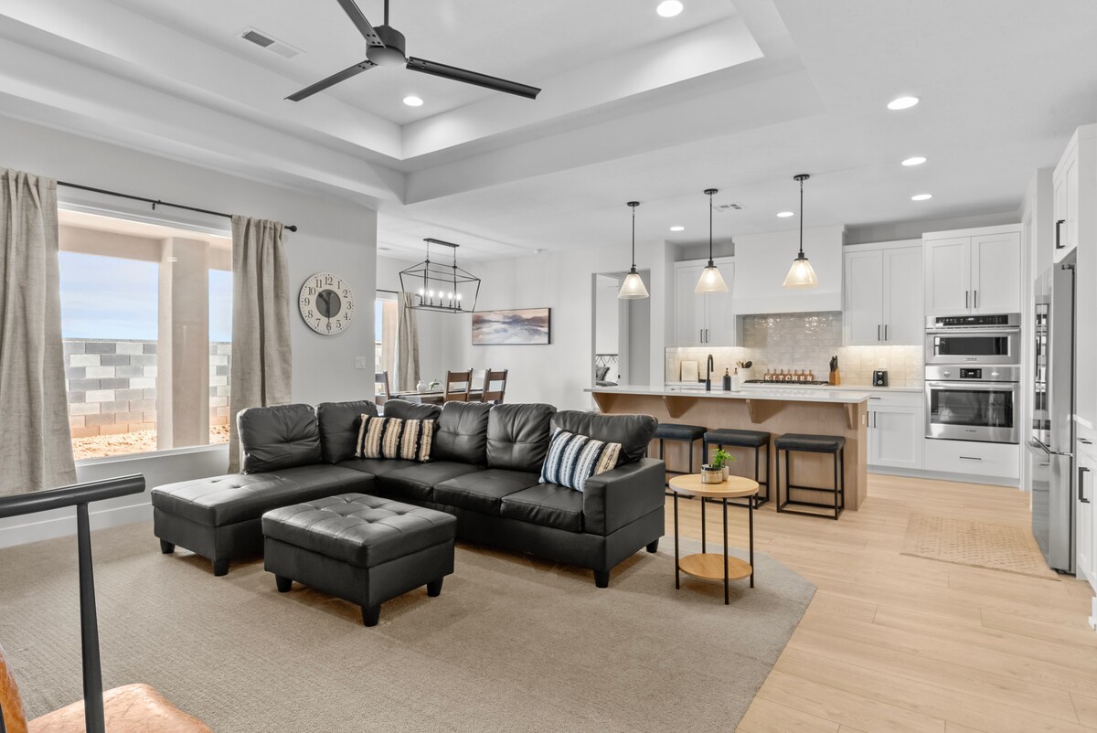 A spacious living area features a modern sectional sofa with accent pillows, a coffee table, and natural light flowing through large windows. The kitchen area includes sleek cabinetry, stainless steel appliances, and a dining table, all set against a backdrop of neutral tones and stylish lighting.
