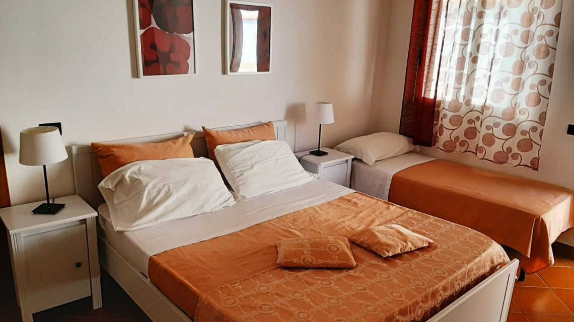 A bright bedroom features a spacious double bed with orange bedding, complemented by a single bed. Two bedside tables with lamps are positioned beside the beds. Natural light enters through a window adorned with patterned curtains, enhancing the room's comfortable layout.