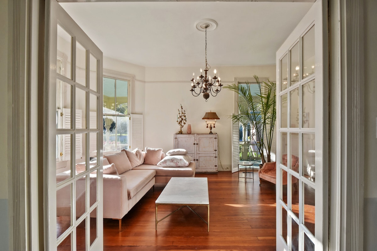 An airy living room is revealed through double French doors, featuring a light pink sectional sofa and a sleek coffee table. Natural light floods the space, highlighting the warm wood flooring and elegant chandelier above. Lush greenery adds a touch of freshness to the atmosphere.