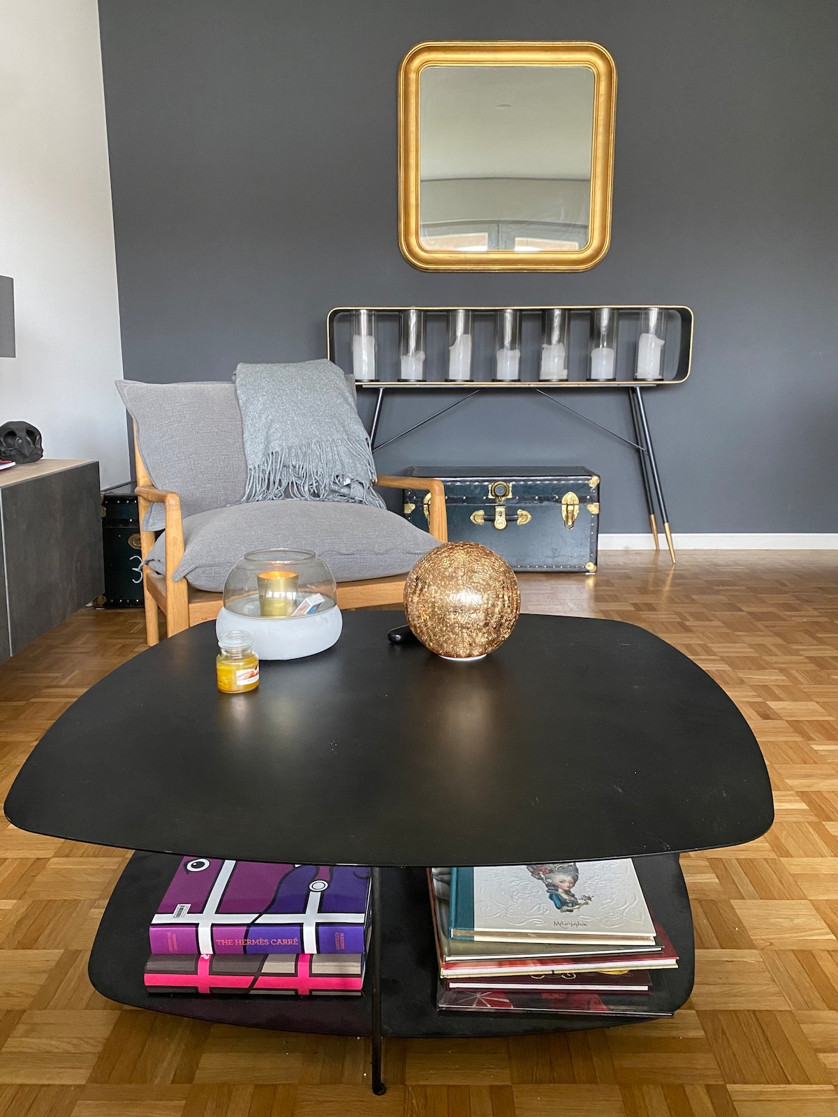 In a modern living area, a light gray chair is positioned beside a black coffee table displaying magazines and decorative items. A circular, golden decorative piece sits atop the table. A mirror with a gold frame is hung on a dark gray wall, adding depth to the space.