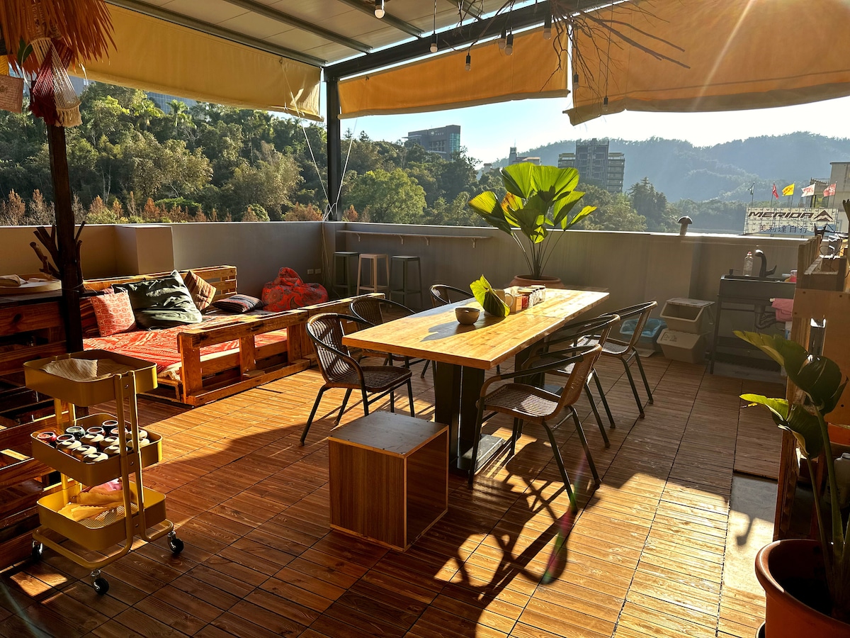 A spacious rooftop area features a wooden dining table surrounded by metal chairs. A cozy seating arrangement is off to one side, complemented by decorative plants. The warm wood flooring and natural light create a relaxed atmosphere, ideal for social gatherings.