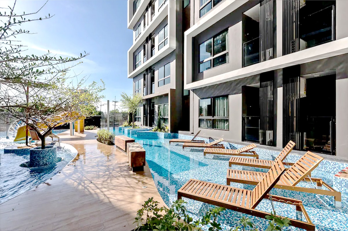 An outdoor pool area is displayed, featuring a bright blue pool with wooden sun loungers positioned along its edge. Lush greenery and decorative trees surround the space, creating an inviting environment for relaxation. A water slide and slides are visible in the background.