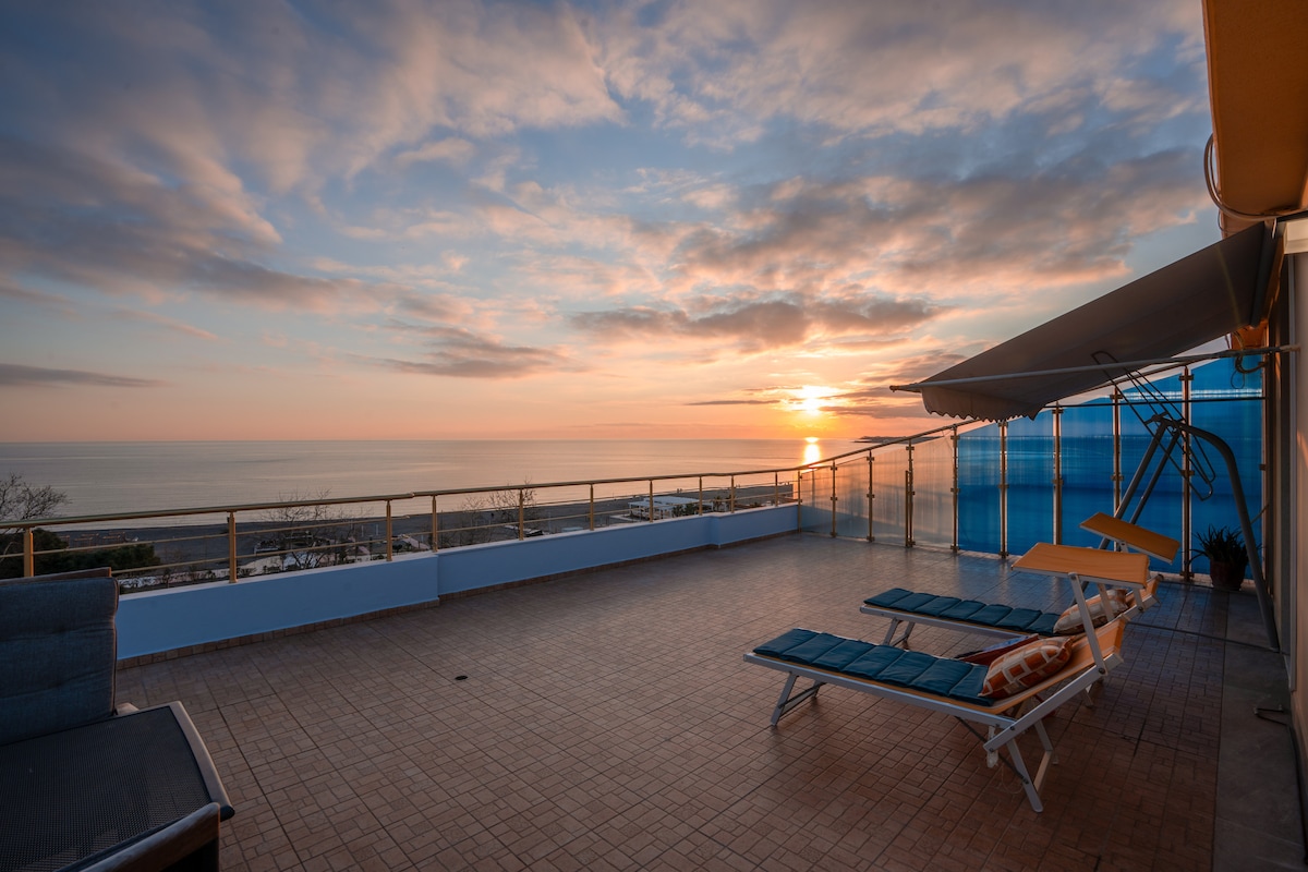 An expansive rooftop terrace offers loungers facing a stunning sunrise over the Adriatic Sea. The sky is filled with soft hues of orange and pink. The terrace features a sleek railing and an awning for shade, creating an inviting space to relax and enjoy the view.