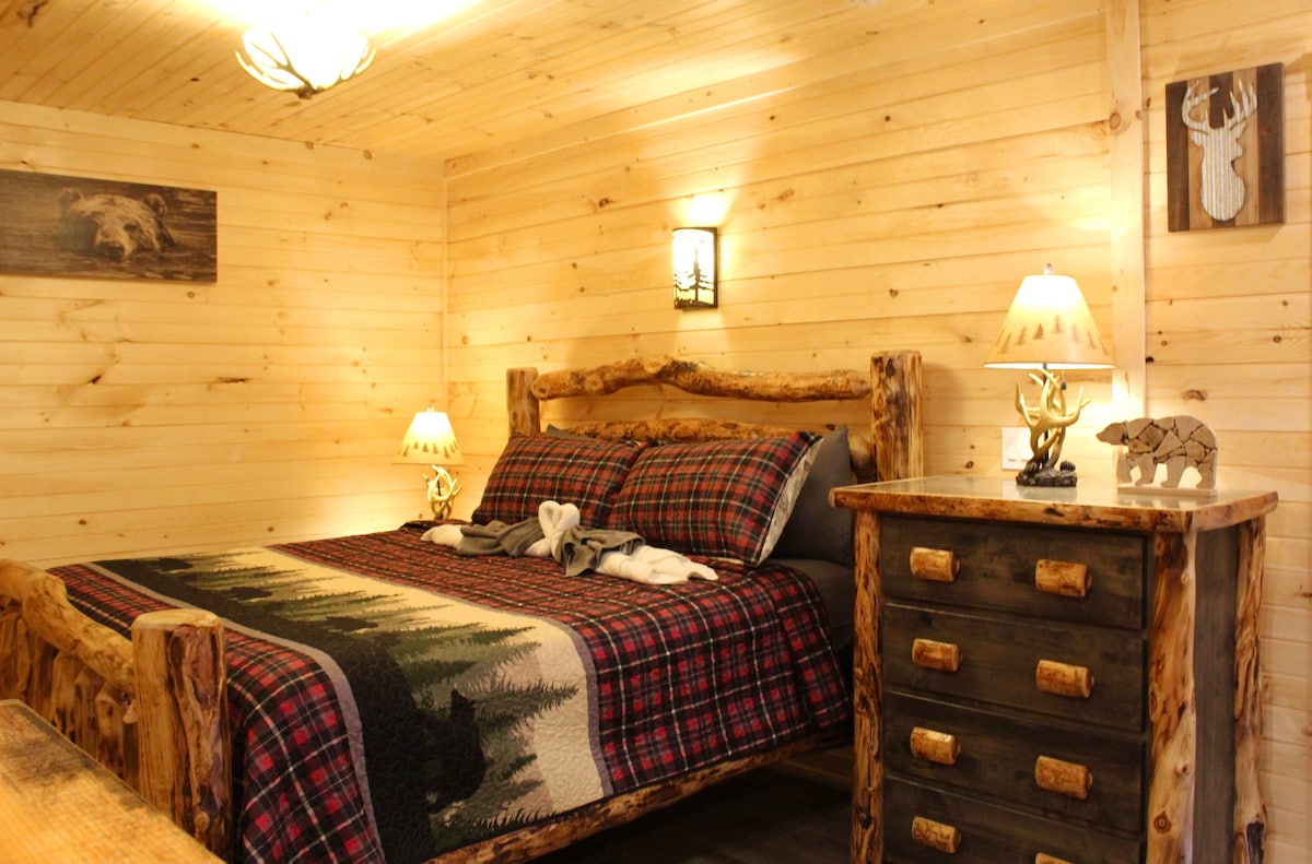 A warm wood-paneled bedroom features a handcrafted log bed adorned with a plaid blanket and cozy throw. Bedside tables with decorative lamps flank the bed, and rustic artwork depicting wildlife accents the walls, creating a charming, rustic atmosphere.