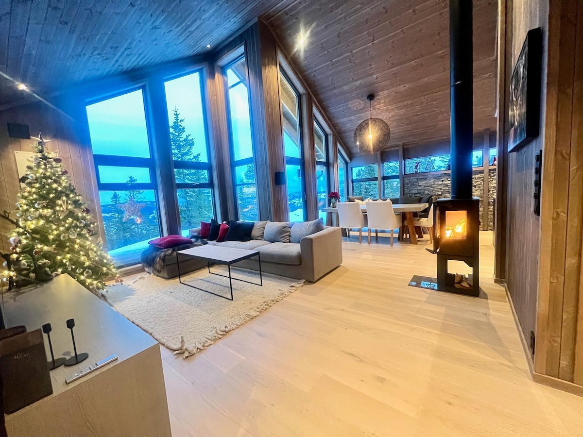 A spacious living area features large windows allowing ample natural light, showcasing scenic views. A comfortable sofa is positioned on a soft area rug, accompanied by decorative pillows. A dining table is visible in the background, and a wood-burning stove adds warmth to the environment.