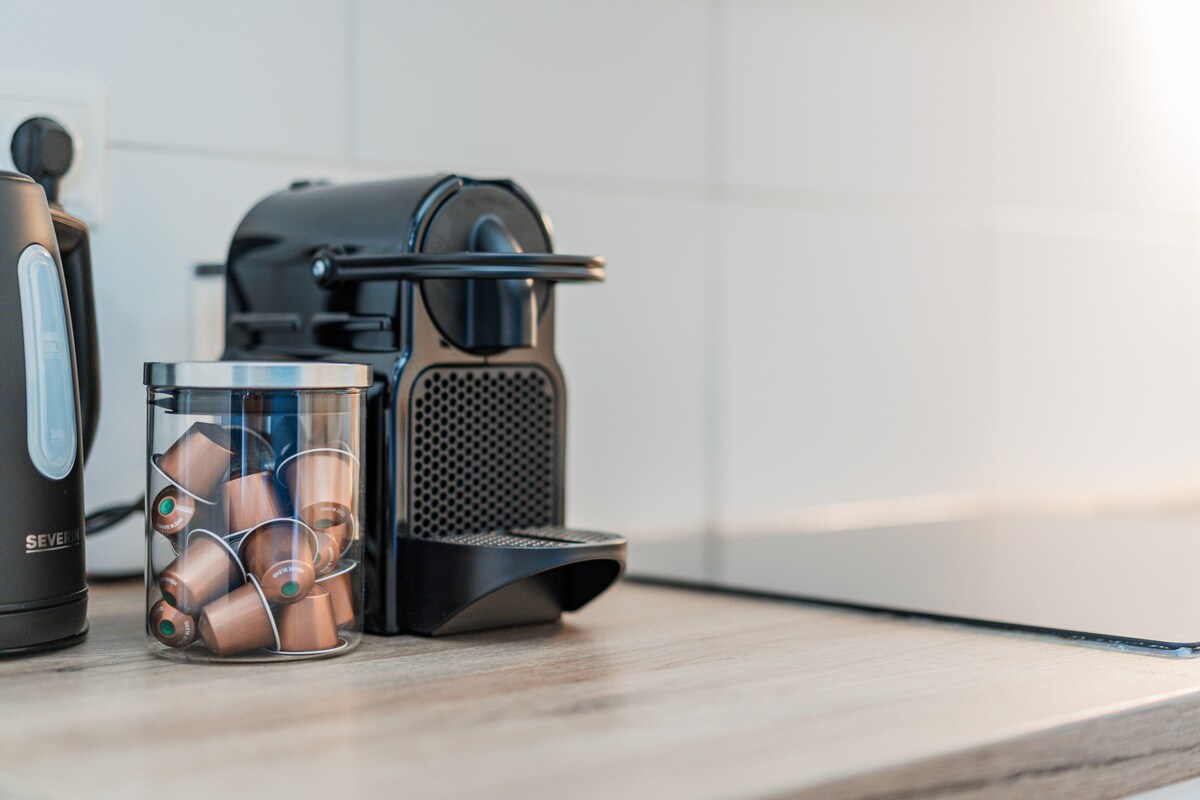 A minimalist kitchen counter is displayed, featuring a sleek coffee machine beside a glass container filled with coffee capsules. A kettle is positioned nearby, and the smooth wooden surface contrasts with the modern appliances.