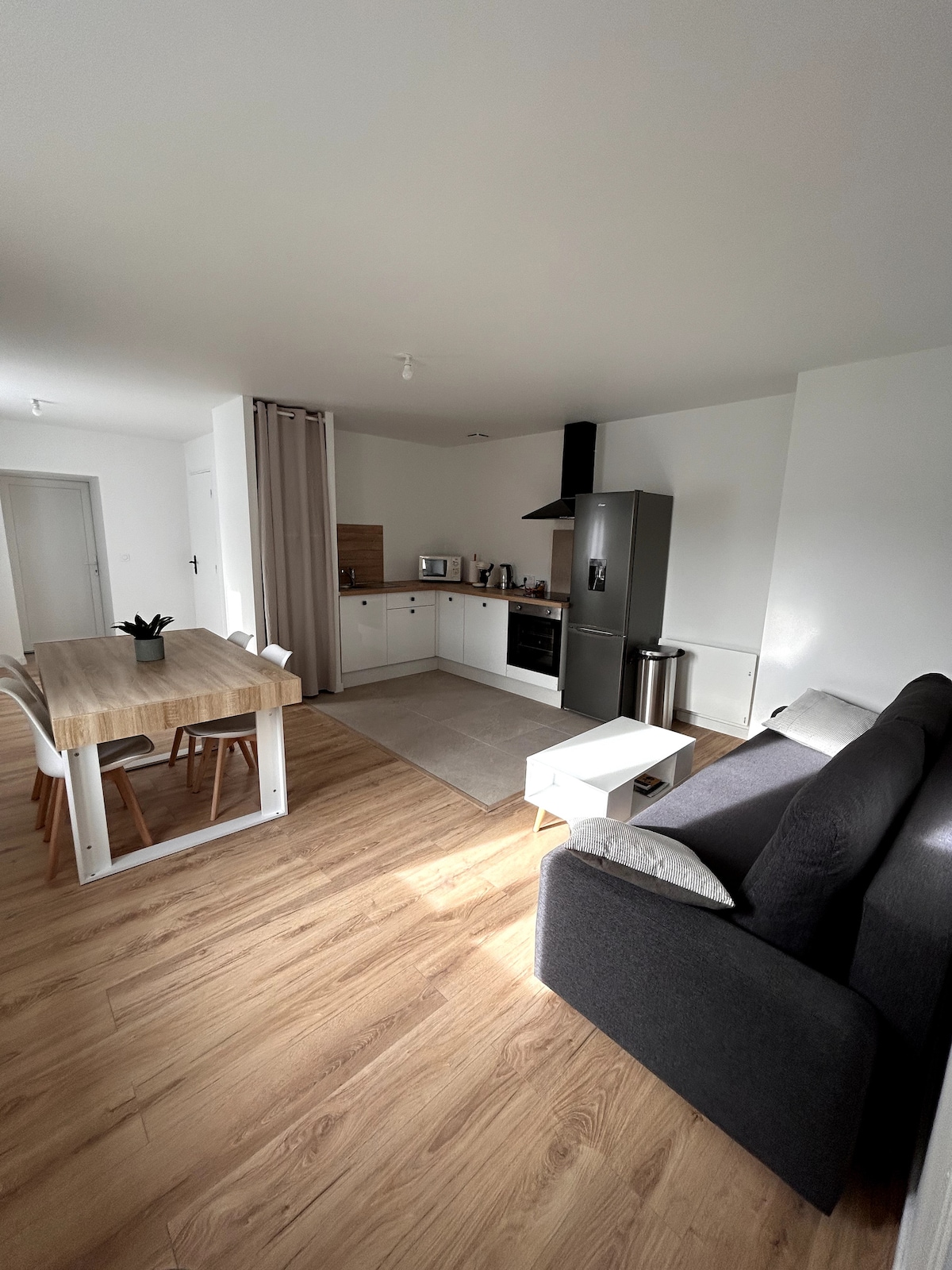 An open-concept living space features a modern kitchen equipped with essential appliances, including a refrigerator and microwave. A dining table with wooden accents is positioned next to the kitchen. A comfortable dark sofa is arranged in the living area, surrounded by natural light.