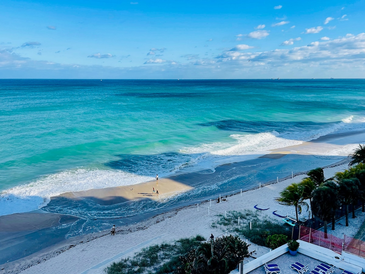 A serene beach view captures gentle waves meeting the shore, with golden sand and a turquoise sea under a bright blue sky. A few people can be seen enjoying the water, while palm trees frame the landscape, adding to the coastal atmosphere.