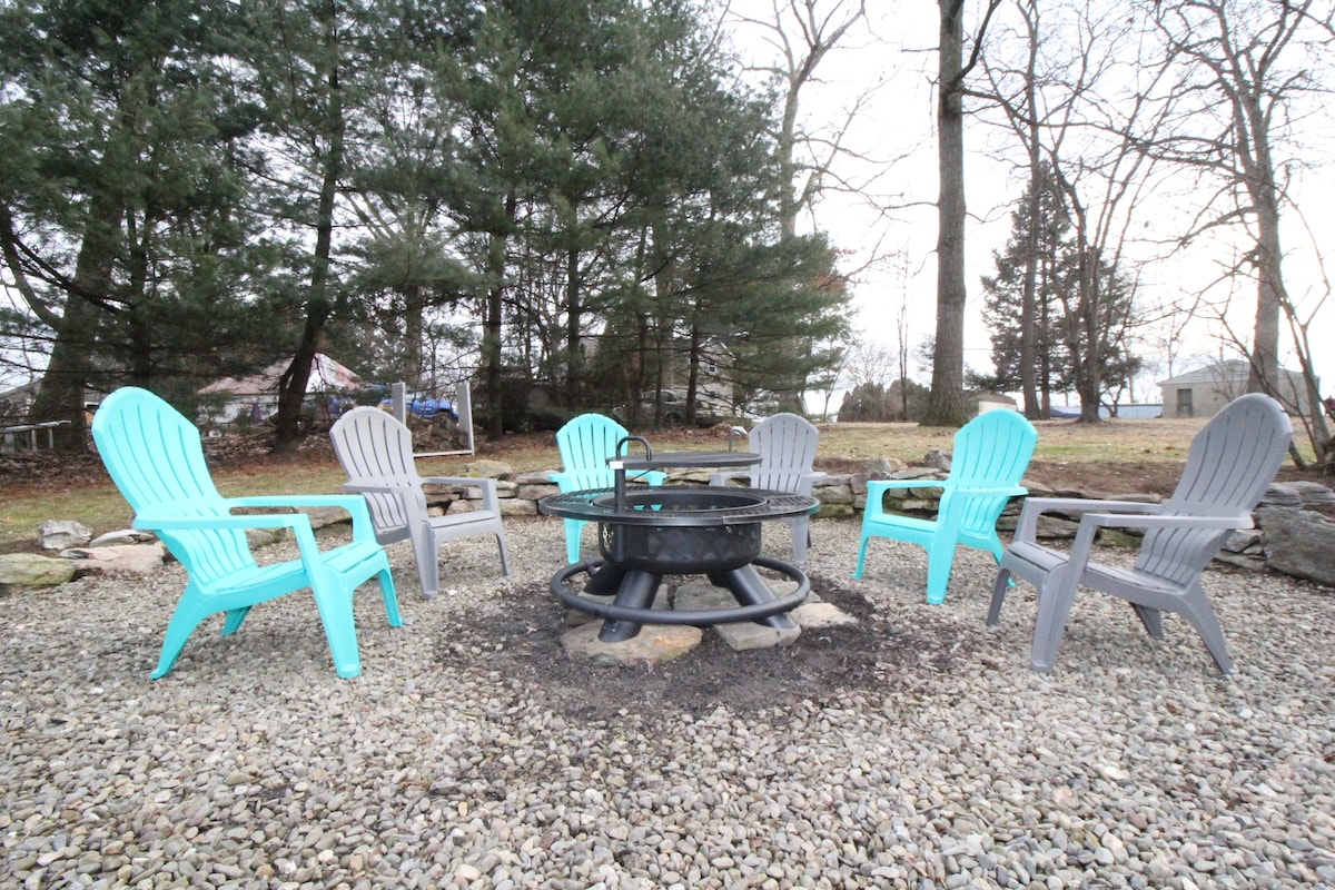 A fire pit area features a circular arrangement of six outdoor chairs, three in blue and three in gray. Surrounding the space, gravel and natural landscaping create a rustic outdoor setting, with trees providing a serene backdrop.