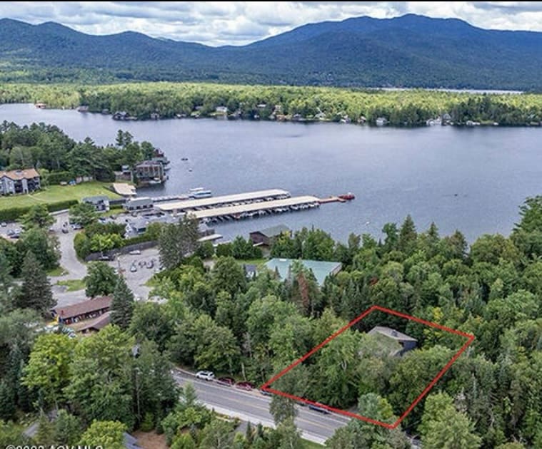 An aerial view captures a serene landscape featuring a lush forest surrounding a contemporary home, outlined in red. The nearby lake glimmers under a cloudy sky, while boat docks and a marina are visible along the water's edge, showcasing the area's outdoor appeal.
