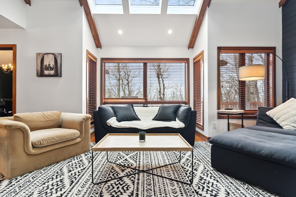 A stylish living area is presented with large windows allowing natural light to fill the space. A cozy sofa and an armchair are arranged around a modern coffee table, situated atop a patterned rug. Warm wood accents complement the overall design.