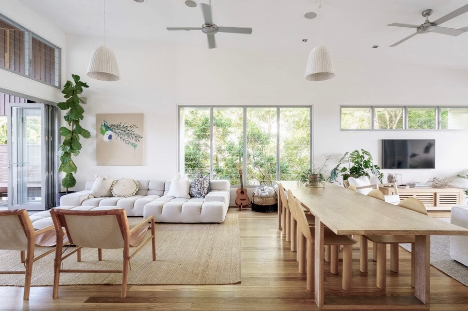 An open plan living area is shown, featuring spacious seating arrangements, including a light-colored sectional sofa and two wooden chairs. A large wooden dining table for eight is centrally positioned. Natural light fills the room through expansive windows, surrounded by greenery.
