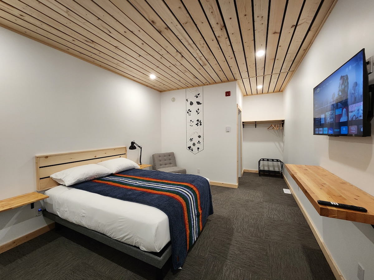 A spacious room features a wooden ceiling and neutral-toned walls. A bed with a colorful blanket is positioned against the wall, complemented by a small seating area. A wall-mounted TV and a desk-like shelf provide additional functionality, while a wardrobe offers storage space.