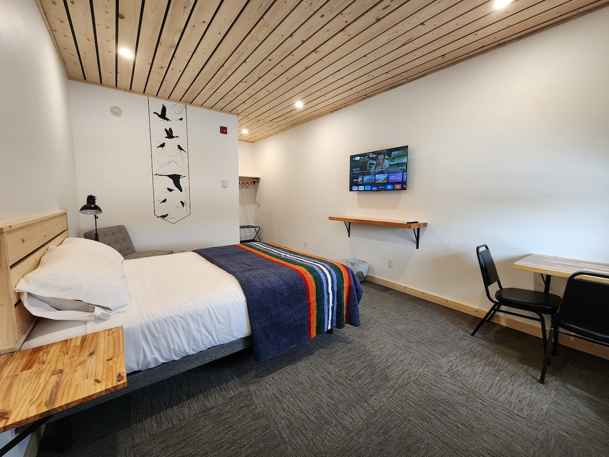 A modern room features a spacious bed with a striped blanket and wooden headboard. A TV is mounted on the wall opposite, with a small table and two chairs nearby. Natural wood accents are visible on the ceiling and side tables.