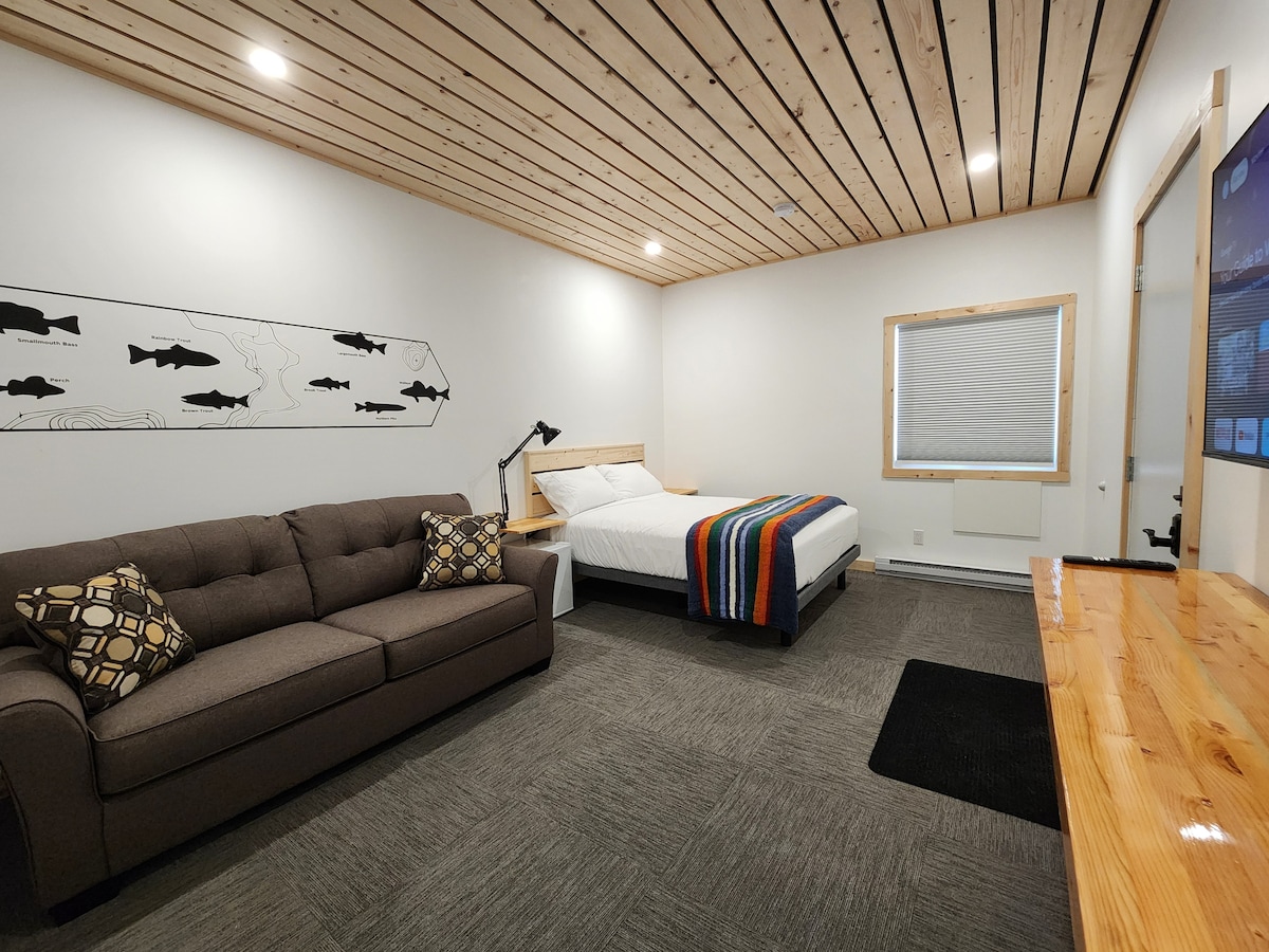 A spacious room features a queen bed adorned with a colorful striped blanket and a cozy sofa, accented by patterned cushions. Natural light enters through a nearby window, while wooden ceiling beams add depth to the modern design.