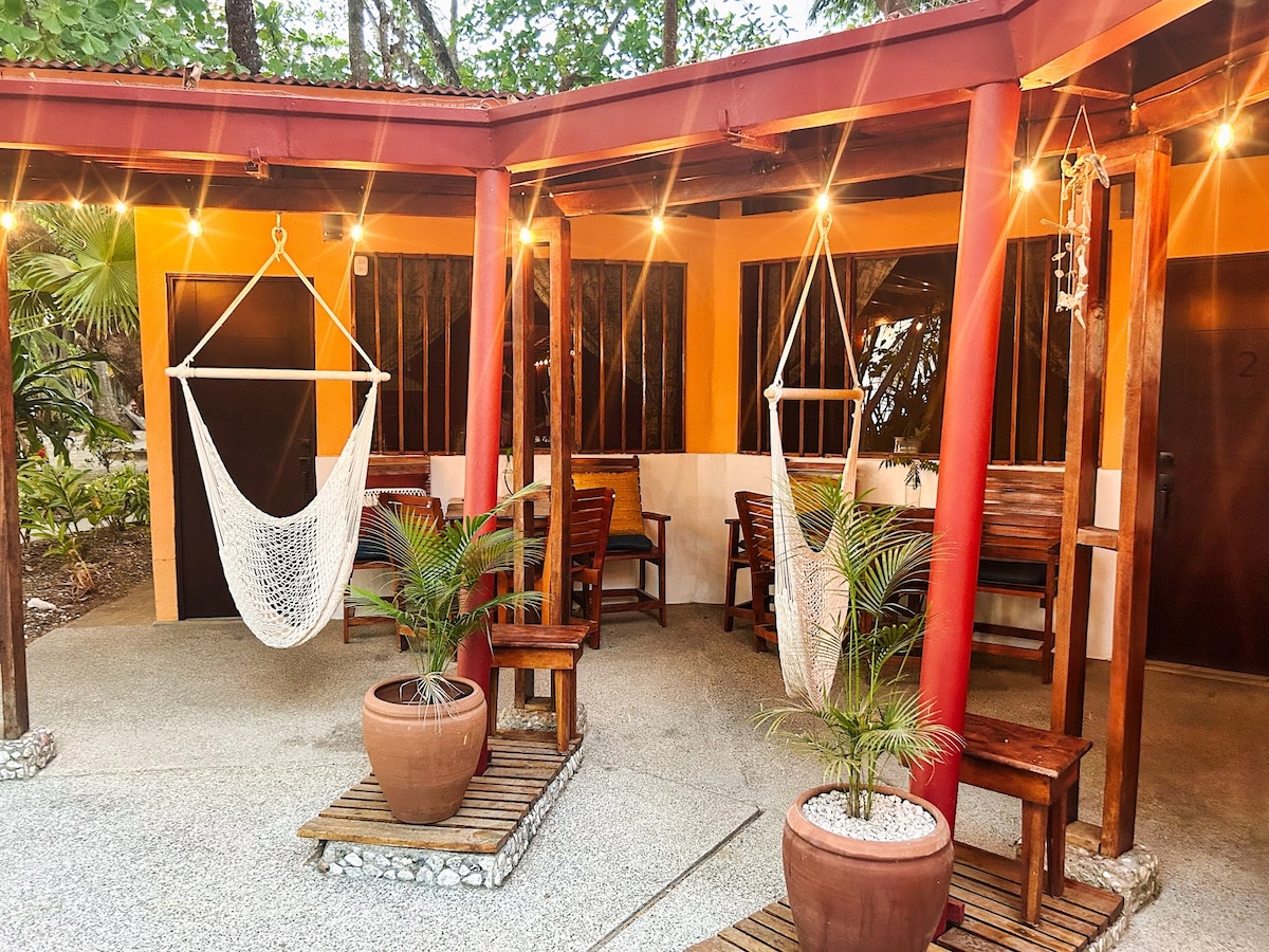A cozy outdoor seating area includes wooden tables and chairs under a rustic canopy. Two hanging hammocks provide relaxation options. Lush greenery surrounds the space, and decorative potted plants add texture. Soft lighting enhances the inviting atmosphere for guests to enjoy.