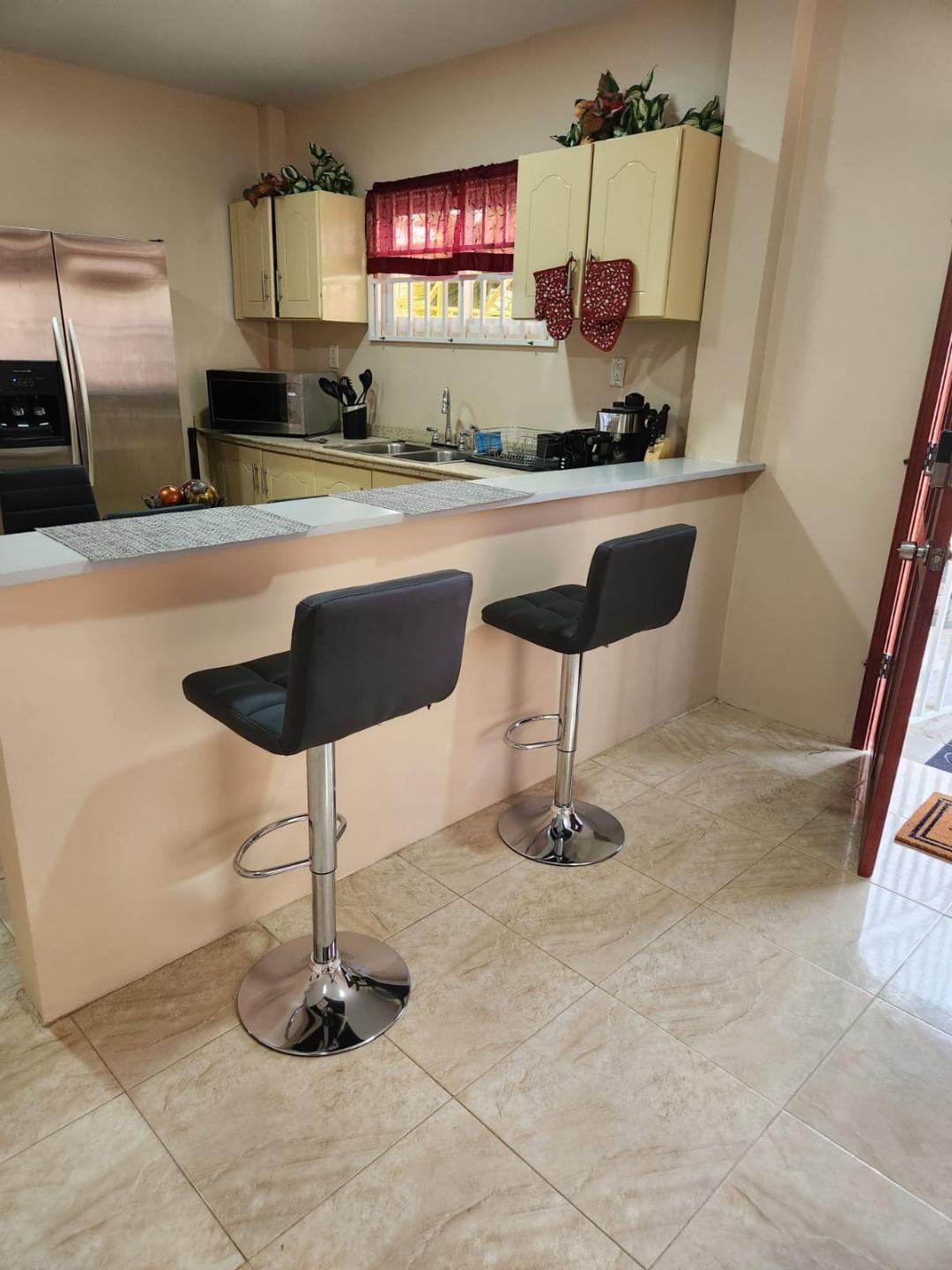 A kitchen area features light-colored cabinetry and stainless steel appliances. Two sleek black bar stools are positioned at a granite countertop, with natural light filtering through a window adorned with red curtains.