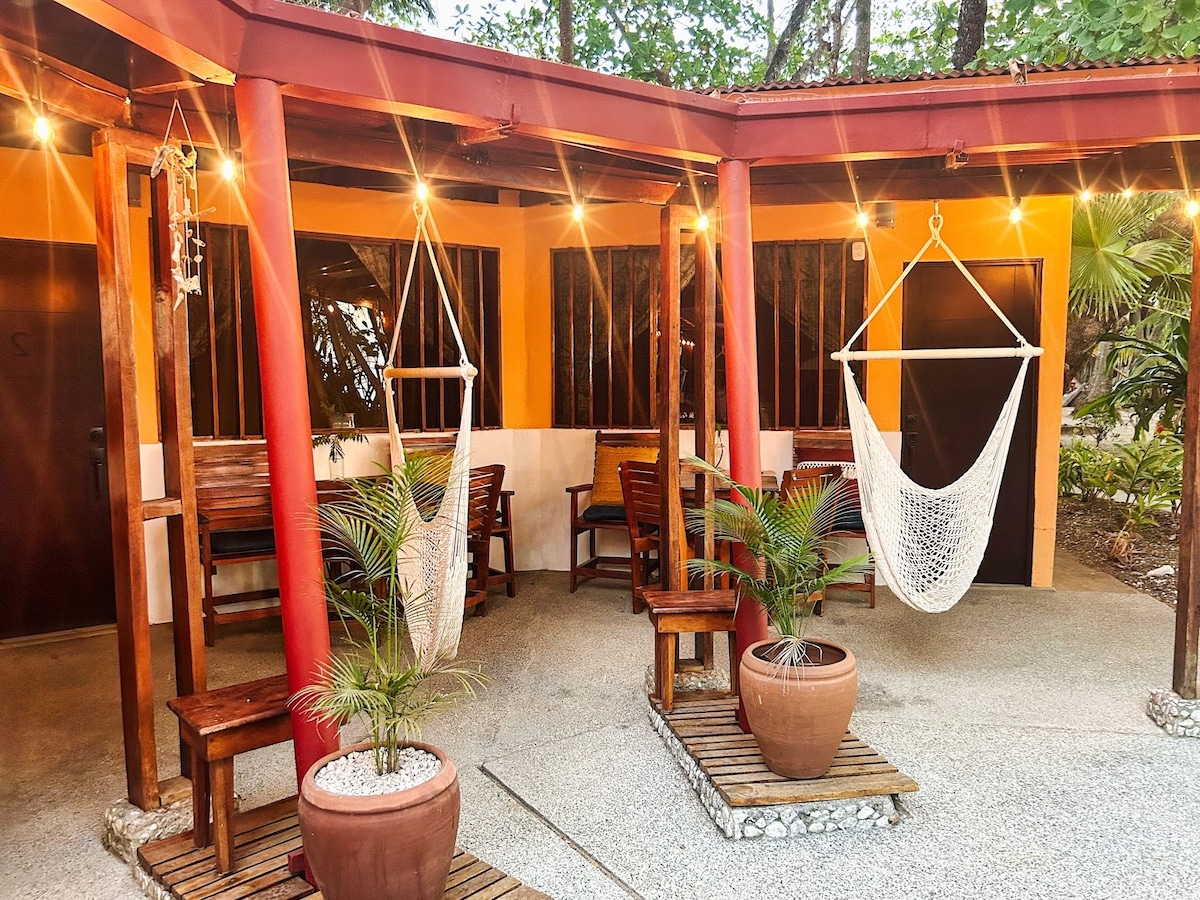 An outdoor seating area is featured, with wooden chairs and a tabletop, surrounded by greenery. Two hanging chairs are suspended nearby, with soft lighting illuminating the space. Potted plants add a touch of nature to the concrete surface, creating a welcoming atmosphere.