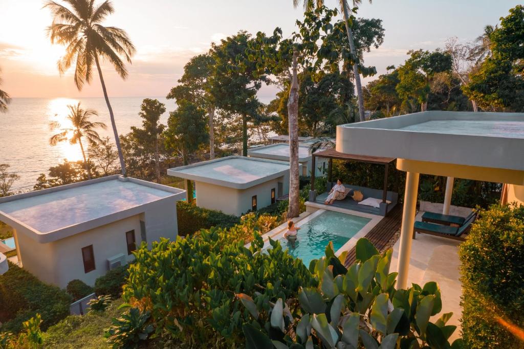 A collection of villas is positioned amidst lush greenery, with a view of the ocean at sunset. An infinity edge pool is featured in the center, accompanied by a relaxation area with comfortable seating. Surrounding palm trees add a tropical ambiance to the scene.