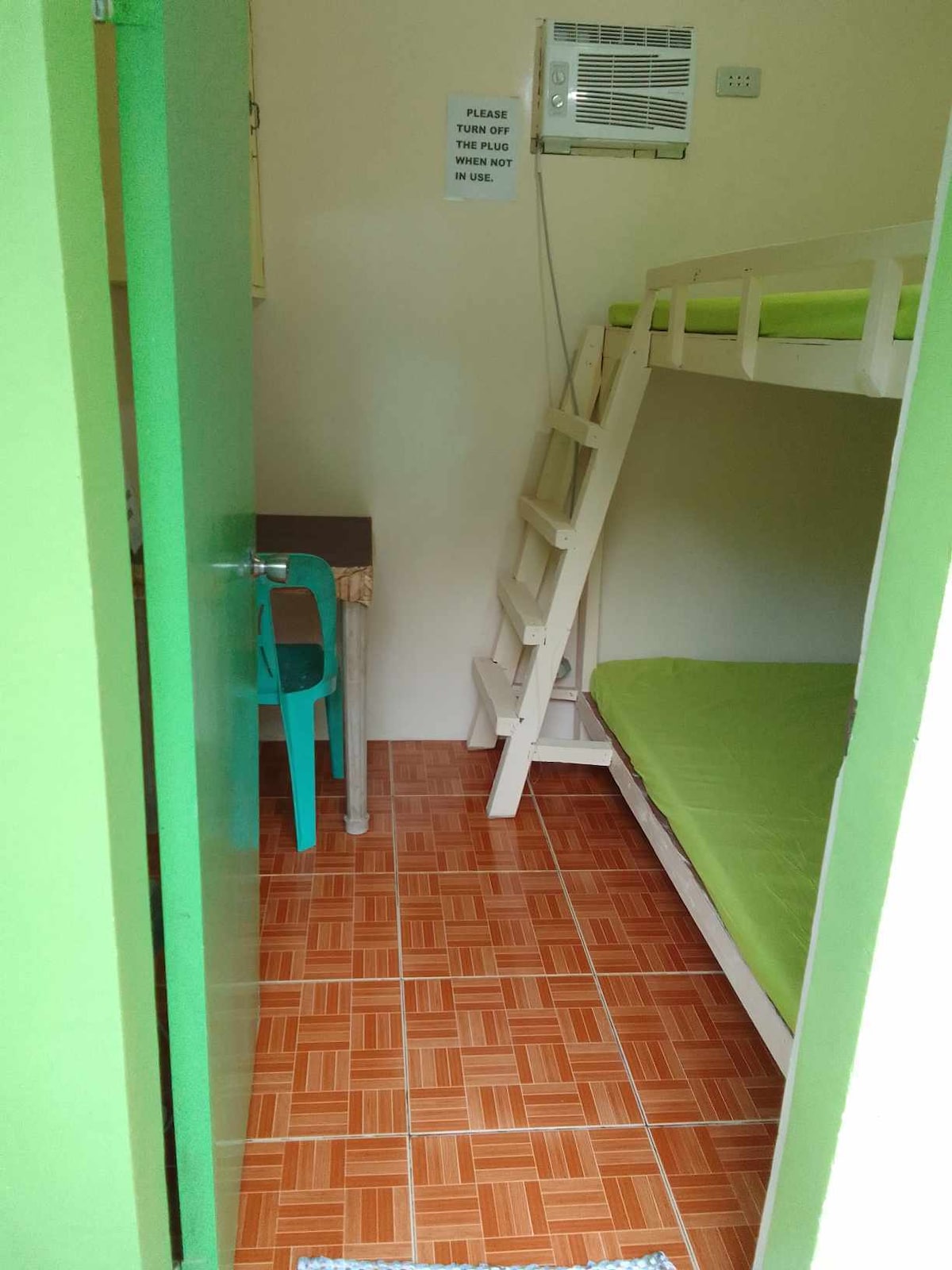 The room features a lofted bunk bed with a green mattress on the top and a lower sleeping area. A small table and a turquoise chair are positioned near the entrance, while tiled flooring adds a warm touch. An air conditioning unit is mounted on the wall.
