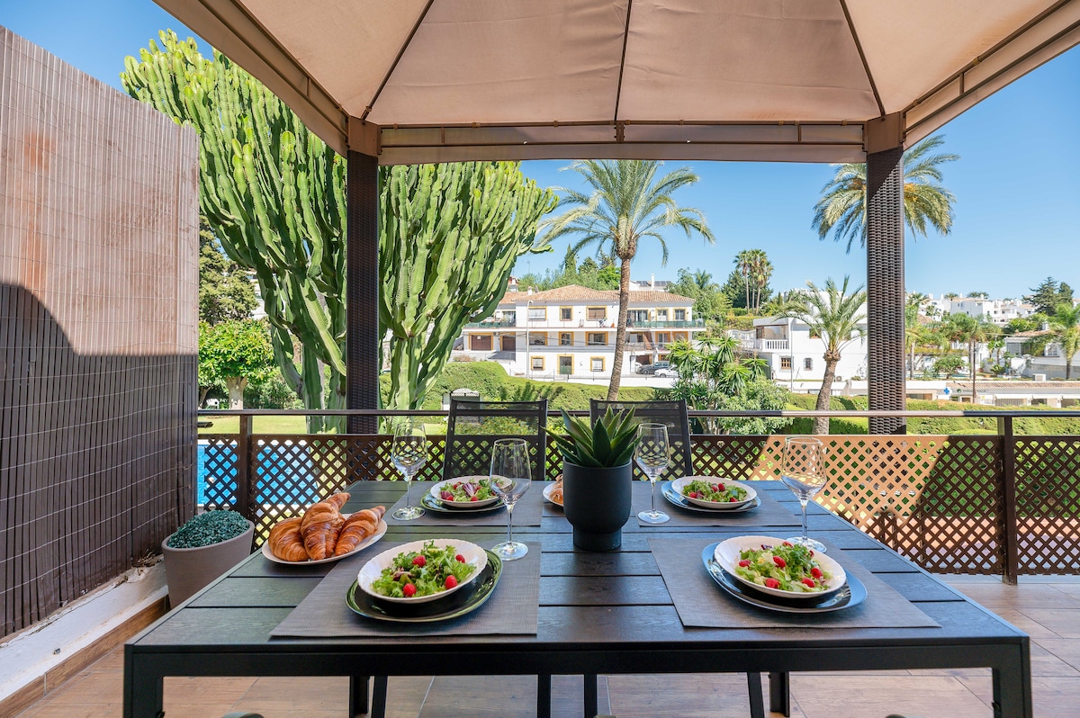 A spacious outdoor terrace features a dining table set for four, complete with plates of salad and croissants. Lush greenery and tall palm trees create a scenic backdrop, enhancing the tranquil atmosphere of the private urbanization.