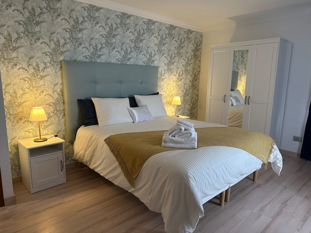 A spacious bedroom is featured, showcasing a padded headboard in soft blue. The bed is dressed in white linens with a light yellow throw. Two bedside lamps provide gentle illumination, and a wardrobe with mirrored doors reflects the serene decor, complemented by a floral wallpaper.