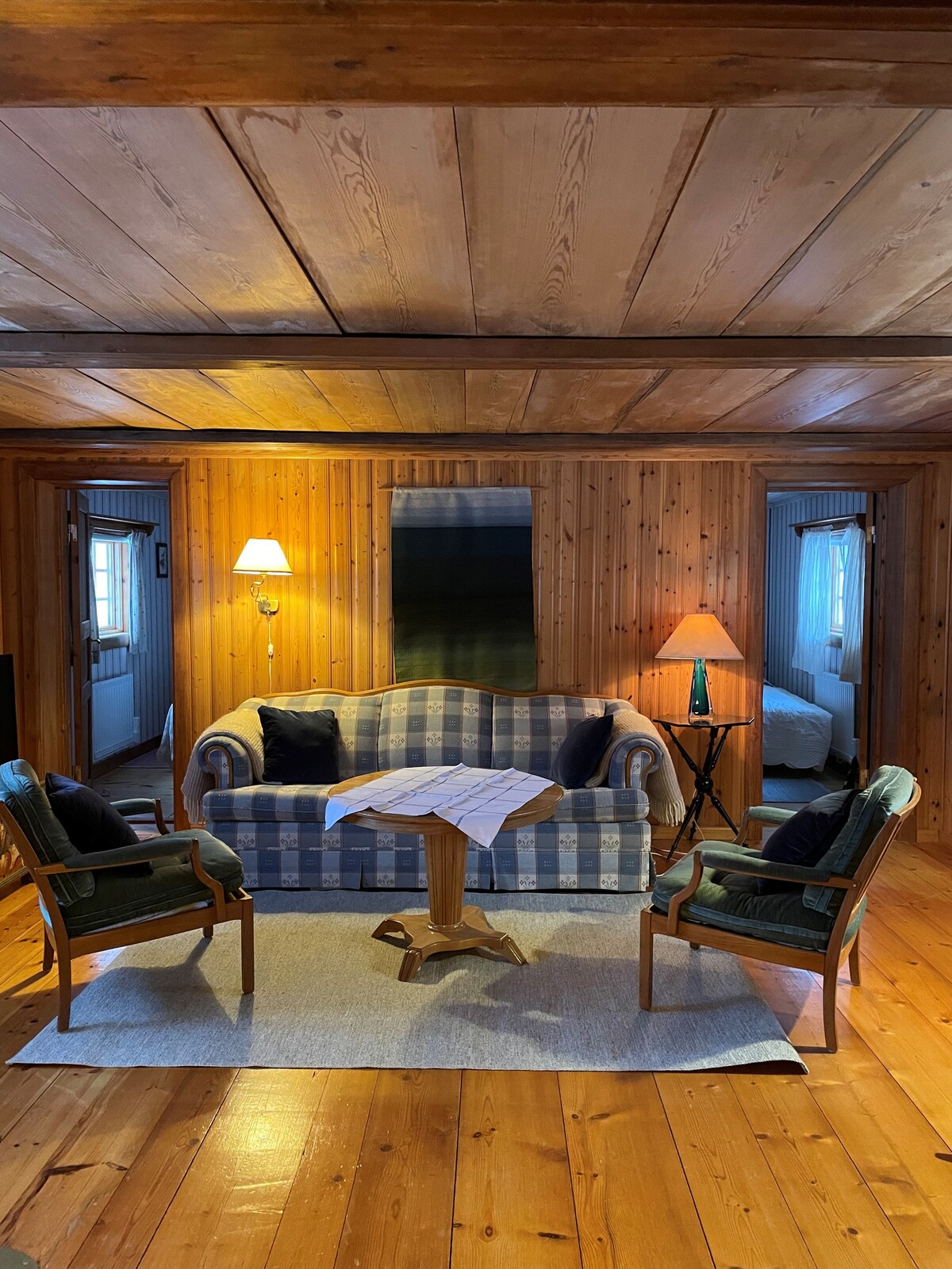 A warm living area is highlighted by a striped sofa positioned in front of a small wooden table. Two armchairs flank the table, with an inviting lamp casting a soft glow. Light wood beams and paneling create a cozy atmosphere.