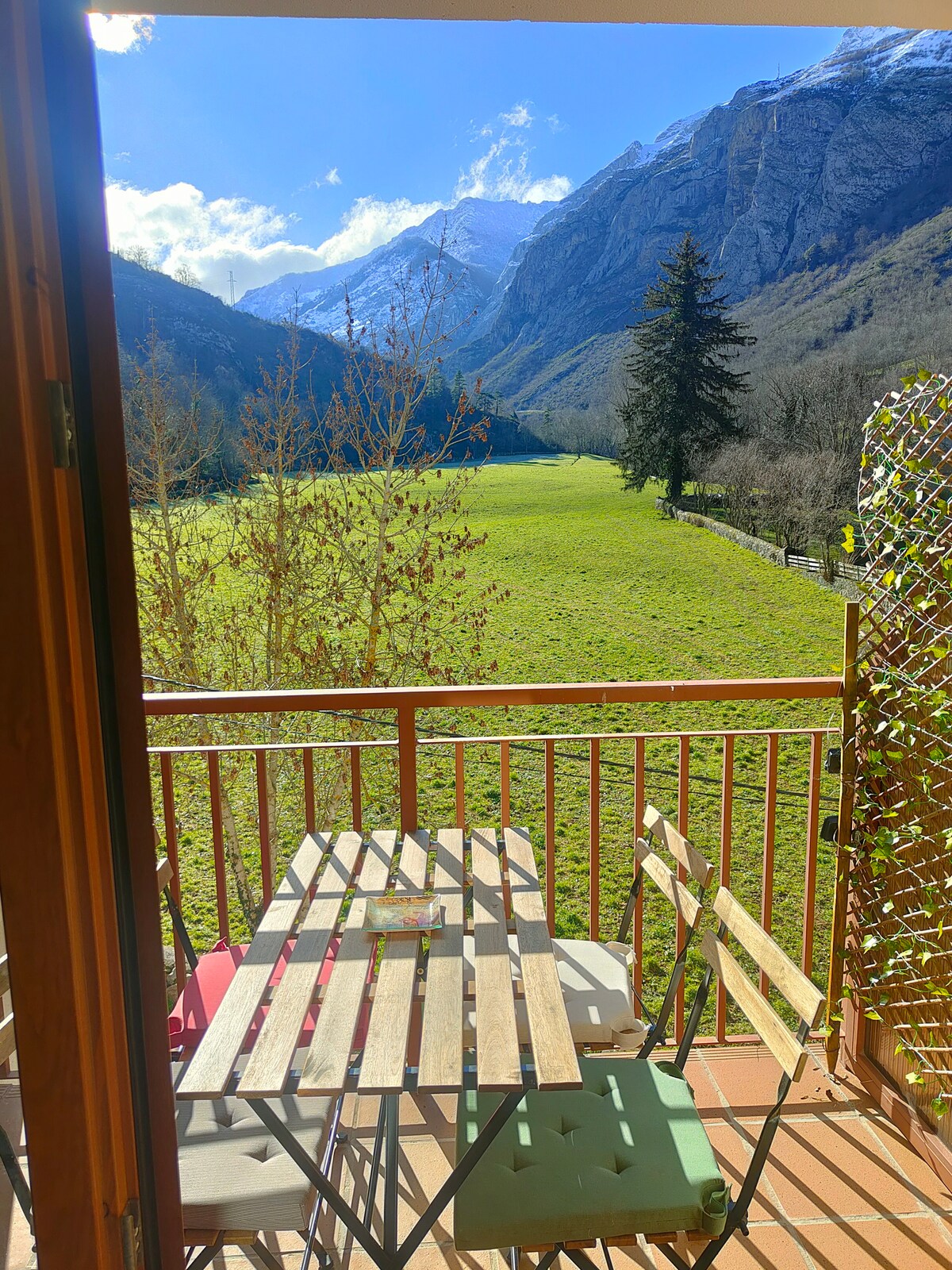A balcony is presented with a wooden table and chairs, accompanied by views of a lush green meadow leading to mountain ranges in the background. Natural light fills the space, highlighting the tranquil outdoor setting.
