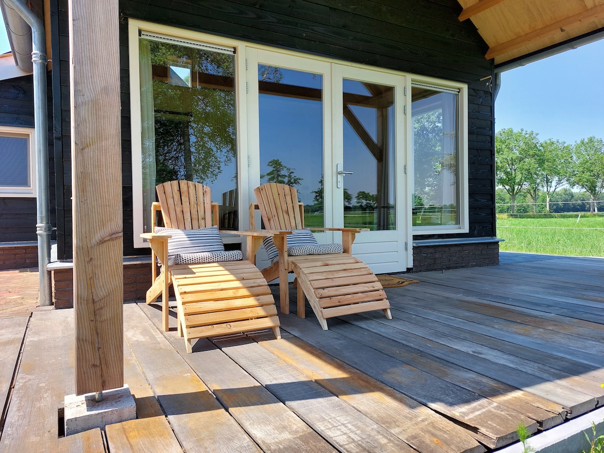 A covered veranda features two wooden lounge chairs positioned for relaxation, with a view of open fields visible through large glass doors. The wooden deck showcases a natural finish, inviting moments of leisure in a tranquil outdoor setting.