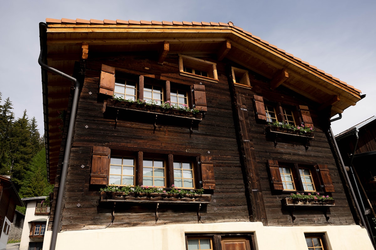 The traditional chalet-style building features a wooden exterior with dark panels and a steep, sloping roof. Wooden shutters frame the windows, which are adorned with flower boxes. Surrounding trees add a natural backdrop to this charming structure.