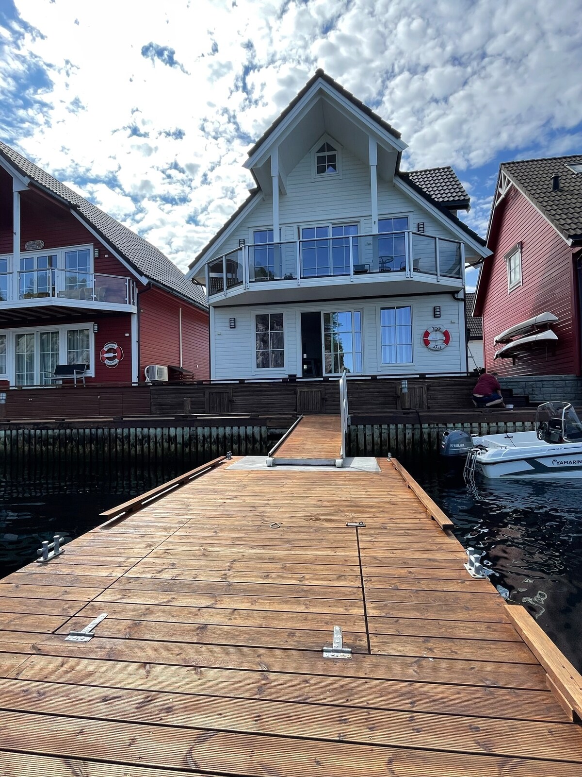 A wooden walkway leads to a spacious two-story rorbu with a light exterior. The structure is flanked by traditional red buildings, and large glass windows provide views of the surrounding area. A boat can be seen docked nearby, with blue sky and clouds reflected on the water.