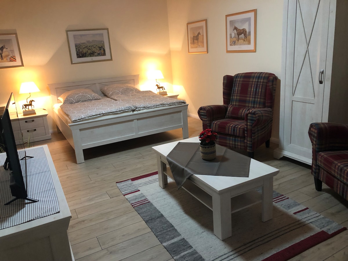 A spacious double room features a light wooden bed with soft bedding, accompanied by two plaid armchairs. A coffee table sits in the center, decorated with a small flower arrangement. The walls display framed horse-themed artwork, and warm lamps provide gentle illumination.