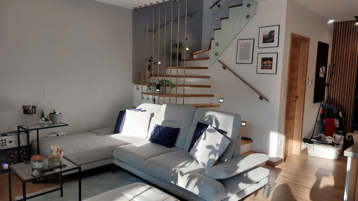 A modern living area features a light gray sectional sofa adorned with dark blue accent pillows. Soft sunlight filters in through a nearby window, highlighting the warm wood tones of the staircase in the background. A small black table is positioned beside the sofa, adding convenience to the space.