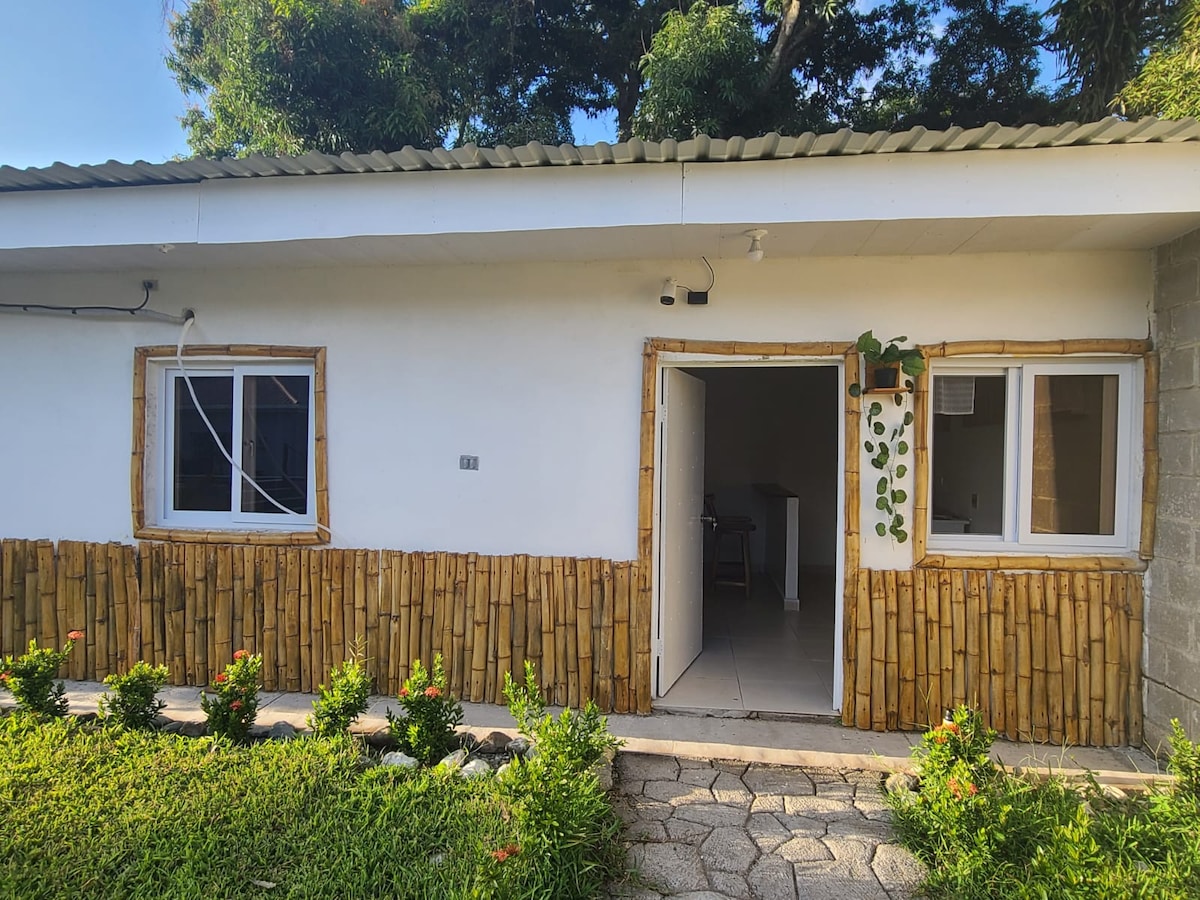 A charming exterior is featured with a white facade accented by natural bamboo elements. Double doors open to reveal a glimpse of the interior. Lush green grass and small flowering plants line the pathway, creating a welcoming entrance to the space.