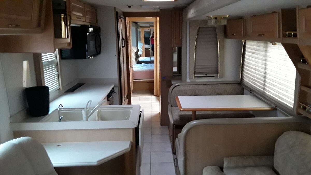 An open interior of a recreational vehicle showcases a well-equipped kitchen area with a double sink and ample counter space. A cozy dining space with a table and specialized seating is visible, while natural light filters through the window treatments.
