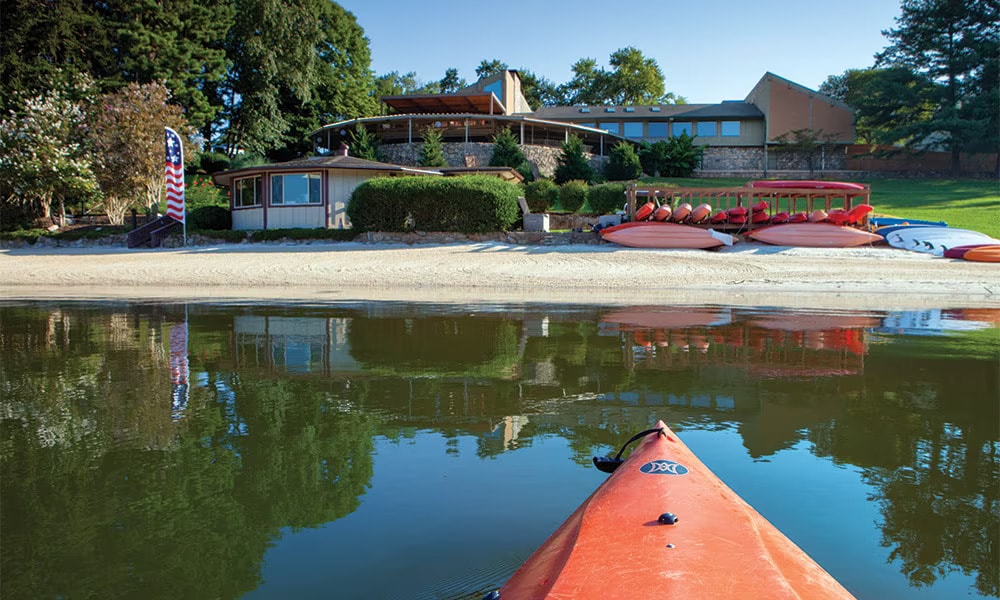The image showcases a shoreline view of a vacation resort, featuring a sandy beach area and several red kayaks lined up along the water. The main building, characterized by a stone façade and large windows, is nestled among green trees, reflecting in the calm lake waters.