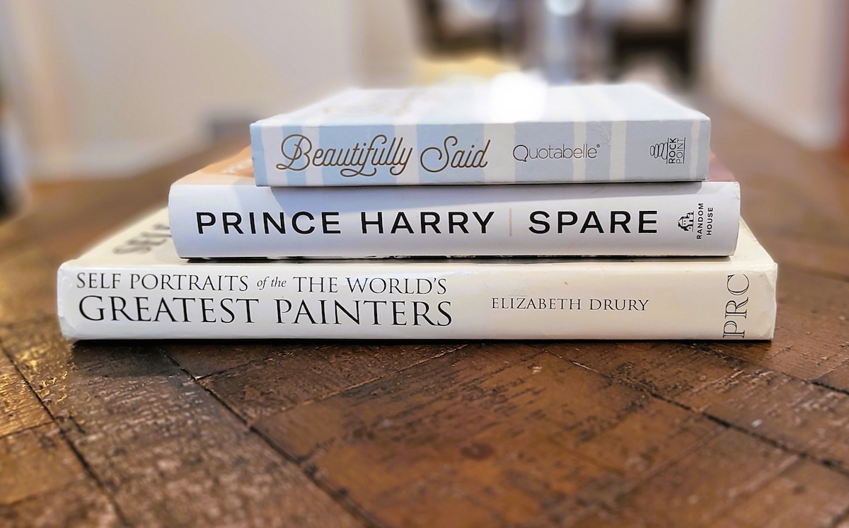 A wooden table displays three stacked books, showcasing their covers. The titles include 'Beautifully Said,' 'Prince Harry Spare,' and 'Self Portraits of the World’s Greatest Painters.' The warm tones of the table enhance the inviting feel of the space.