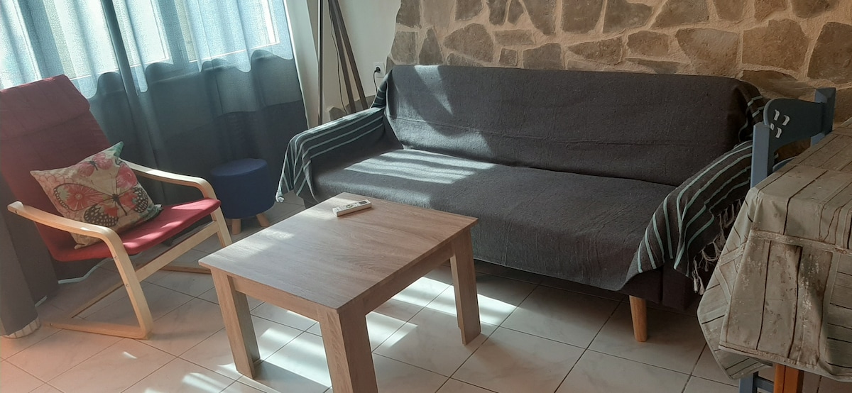 A cozy living area is presented with a comfortable sofa and a wooden coffee table, illuminated by natural light from nearby windows. A red armchair with a decorative pillow adds a pop of color, while a stone wall provides a textured backdrop.