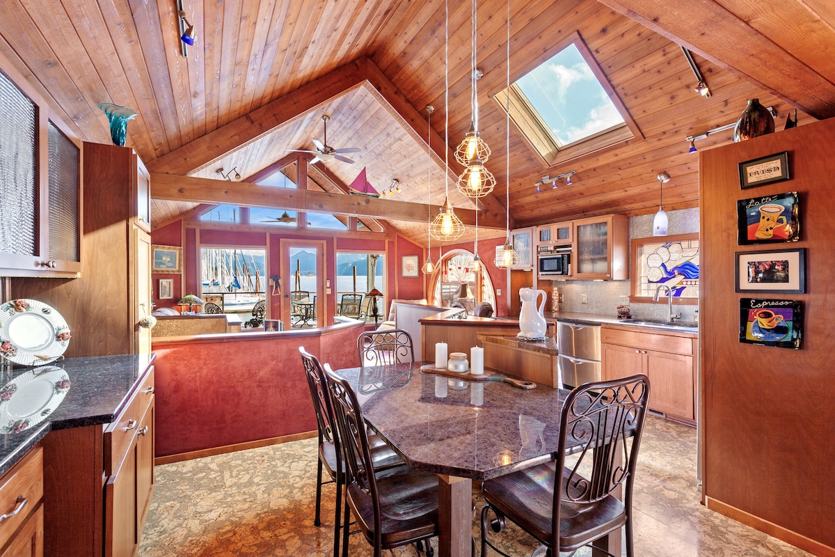 The kitchen features wooden ceilings and warm tones, with a central table surrounded by stylish chairs. Large windows provide natural light and views of the lake, while modern appliances are integrated into the cabinetry, offering a functional and inviting cooking space.