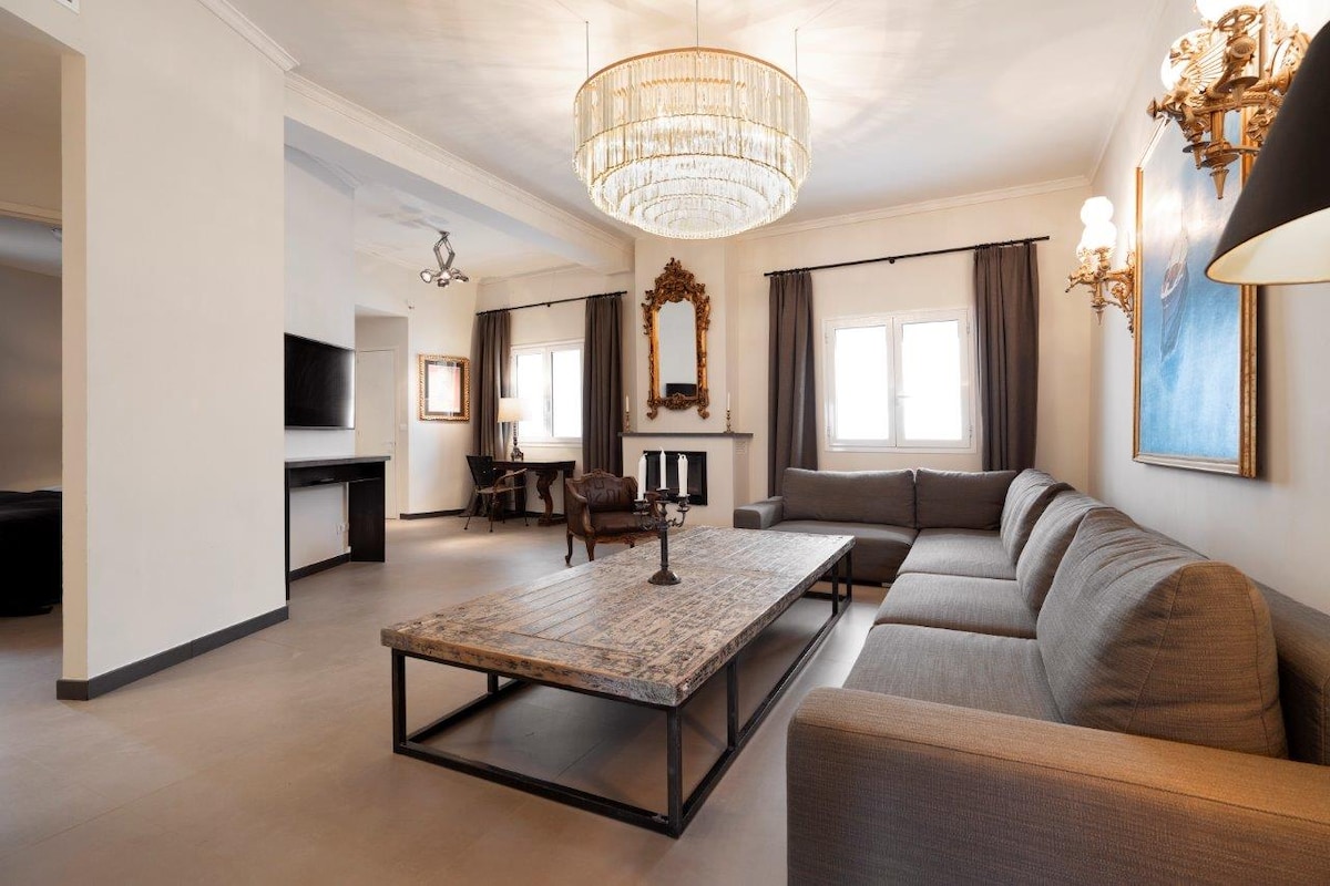 A spacious living area is highlighted by a large coffee table at the center. Soft gray sofas complement the neutral color scheme, while natural light enters through the windows adorned with elegant curtains. A decorative mirror and artwork enhance the ambiance of the room.