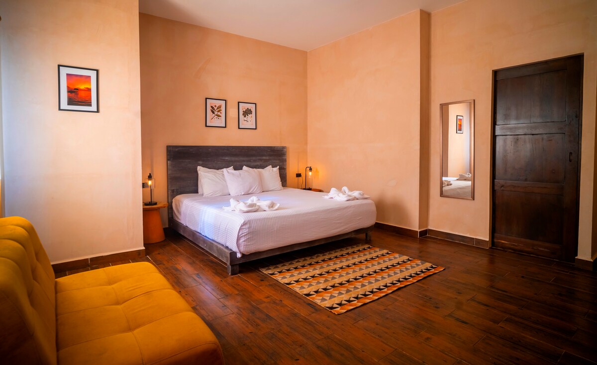 A bright and airy bedroom features a spacious king-sized bed adorned with freshly laundered linens. A cozy sofa is positioned near the window, and framed artwork is displayed on the walls. A mirror reflects the warm tones of the room, enhancing the inviting environment.